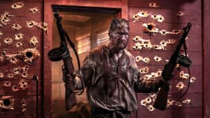 Huvudkaraktären i "Sisu: Road to Revenge" med två skjutvapen i händerna och en massa kulhål bakom sig.