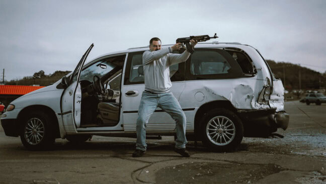 En man stor vid en krockskadad bil och håller ett skjutvapen i händerna.