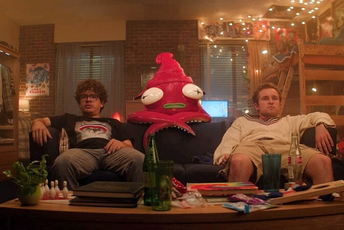 Två snubbar i en soffa, med ett udda och storögt lila monster bakom sig.