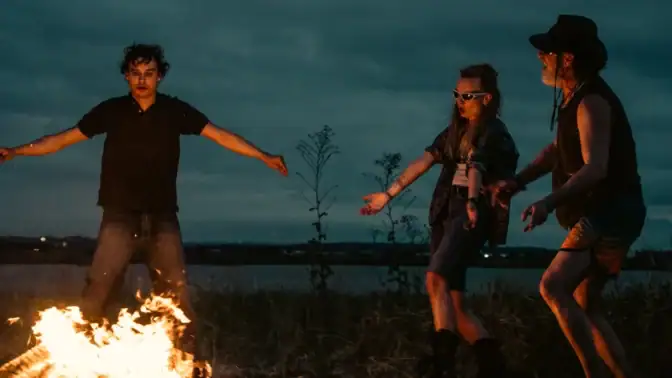 Tre personer dansar runt en lägereld.