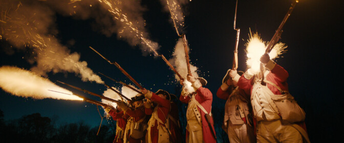 "Historical reenactors in red military uniforms fire muskets in unison at dusk, with bright muzzle flashes lighting up the night sky. The scene suggests a battle setting, possibly from the 18th century."